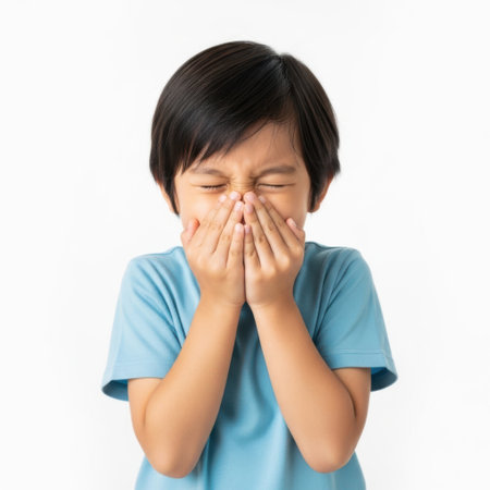 A young child with dark hair, wearing a blue t-shirt, is holding their hands over their nose and mouth, eyes closed, appearing to sneeze or cough.の素材