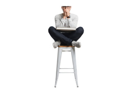 A young person in casual clothing sits cross-legged on a tall stool, engrossed in reading a book, with a thoughtful expression.の素材