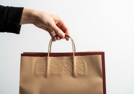 A person's hand holds the handles of a brown recycled paper shopping bag against a plain white backdrop.の素材