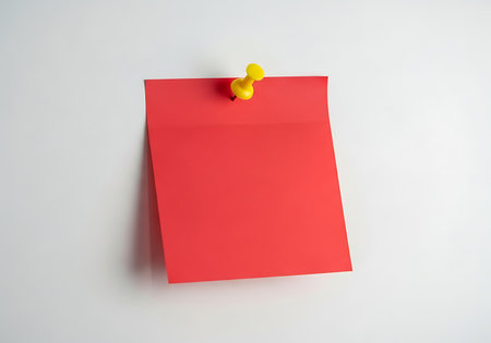 A vibrant red note card, commonly used for reminders, is securely attached to a clean white wall. A bright yellow thumbtack pierces the top corner, holding the note in place. The simple composition highlights the bold color.の素材