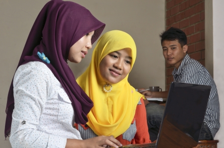 Two young Muslim ladies with laptop discussing, while a young man looking in the backgroundの写真素材