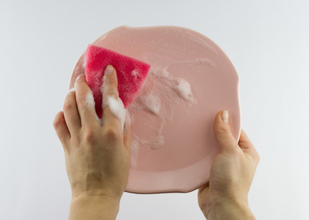 Housekeeping concept. Female hand with red sponge washing pink plate isolated on grey backgroundの写真素材