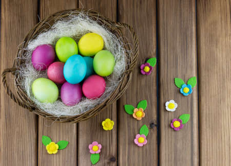 Top view of basket with colorful painted eggs and sugar flowers on wooden background