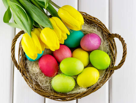 Top view of yellow tulips with basket full of colorful painted Easter eggs on white backgroundの写真素材