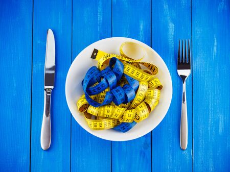 Healthy lifestyle concept. Top view of white plate full of measuring tape with silver fork and knife on blue wooden backgroundの写真素材