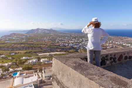 Young redhead woman satnding and watching the traditional villages at sunny windy day. Lifestyle and travel concept. Santorini, Greece.の写真素材