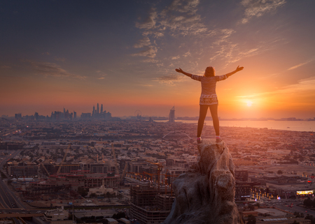 Young woman standing on the rock in a beautiful open sunset scene on city with her arms raised up in the air with confident feeling. Success and freedom concept.の写真素材