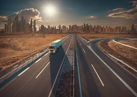 White truck on straight highway towards the sun and beautiful skyscrapers of cityscape at sunset. Transportation concept.の写真素材