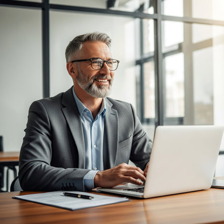 Cheerful mature businessman in eyeglasses using laptop while working in officeの素材