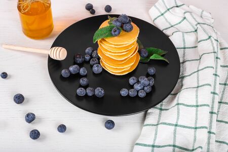 image shows a homemade fluffy pancake with blueberry and mint on the top; situation is decorated with white wooden table and honey glass; black plate with pancakesの写真素材