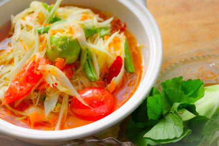 Delicious papaya salad on wooden table and some fresh vegetables on dish. This is famous local thai...
