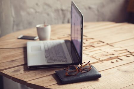 Wooden table with laptop, glasses, wallet and cup of coffeeの写真素材
