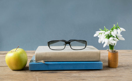 Creative book arrangement with eyeglasses, apple and snowdrops on wooden table.