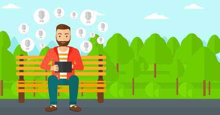 A hipster man with the beard sitting on a bench in the park and holding a tablet computer with many avatar icons above vector flat design illustration. Horizontal layout.のイラスト素材