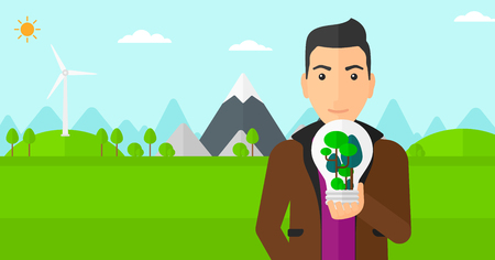 A man standing on a background with wind turbins and holding in hands a big lightbulb with trees inside vector flat design illustration. Horizontal layout.のイラスト素材