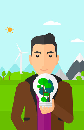 A man standing on a background with wind turbins and holding in hands a big lightbulb with trees inside vector flat design illustration. Vertical layout.のイラスト素材