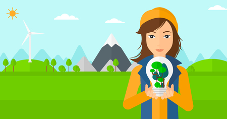 A woman standing on a background with wind turbins and holding in hands a big lightbulb with trees inside vector flat design illustration. Horizontal layout.のイラスト素材