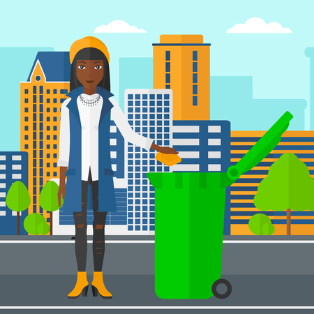 An african-american woman throwing a trash into a green bin on a city background vector flat design illustration. Square layout.のイラスト素材