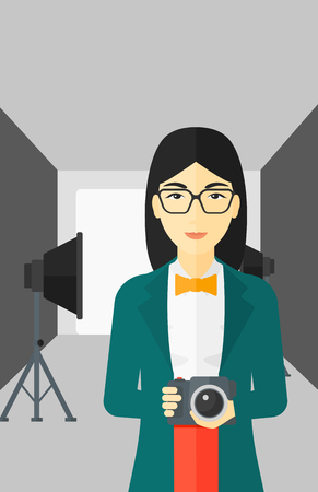 An asian woman holding a camera on the background of photo studio with lighting equipment vector flat design illustration. Vertical layout.のイラスト素材