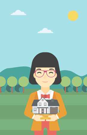 An asian young woman holding house model in hands on the background of mountains. Real estate agent with house model in hands. Vector flat design illustration. Vertical layout.のイラスト素材