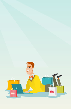Young caucasian man using a laptop for online shopping. Smiling man lying with a laptop and shopping bags around him. Happy man doing online shopping. Vector flat design illustration. Vertical layout.のイラスト素材