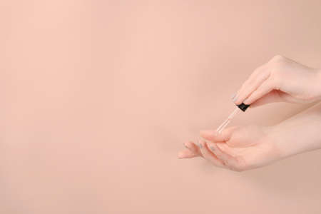 Woman with pastel manicure used serum dropping on her hand on beige background. Skin care and beauty concept. Front view with copy spaceの写真素材
