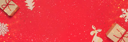 Baner with craft gift box with red ribbon as a presenf for Christmas, new year on red background with baubles, lights and confety. Flat lay copy spaceの写真素材