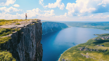 A solitary figure stands on a dramatic cliff edge, gazing over a shimmering lake framed by verdant hills and a vibrant sky. A perfect scene for adventure and peace.の素材