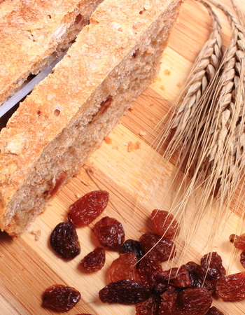 Slicing fresh baked wholemeal bread, ears of wheat and heap of raisins lying on cutting board, concept for healthy eatingの写真素材