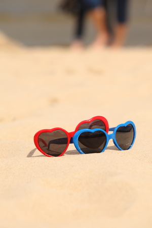 Red and blue funny sunglasses in the shape of a heart lying on the sand at the beach, summer timeの写真素材