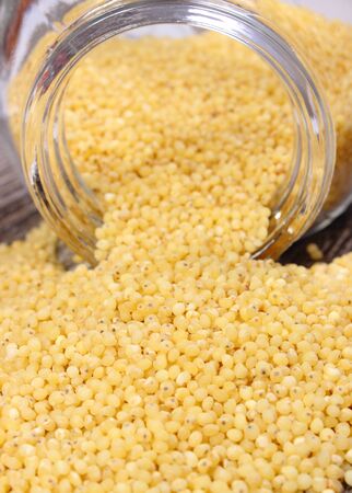 Heap of yellow millet groats spilling out of glass jar on wooden background, concept for healthy...