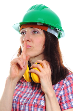 Female construction worker wearing green helmet and protective headphones, holding finger to lips and showing silence sign, safety at work and ear protection. White backgroundの写真素材