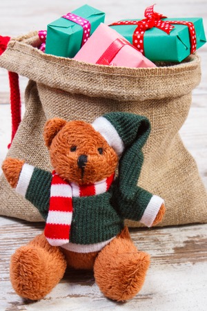 Funny teddy bear and wrapped colorful gifts for Christmas, birthday, valentines or other celebration in jute bag on old wooden white tableの写真素材