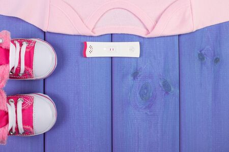 Pregnancy test with positive result and clothing for newborn on purple boards, concept of extending family and expecting for baby, copy space for text or inscriptionの写真素材