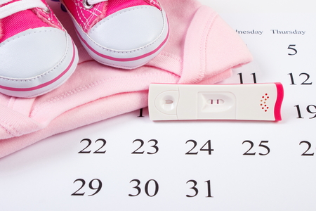 Pregnancy test with positive result and clothing for newborn on calendar, baby shoes, bodysuits, concept of extending family and expecting for babyの写真素材