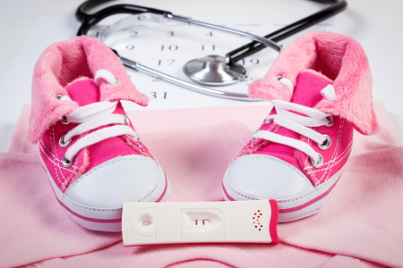 Pregnancy test with positive result, clothing for newborn and stethoscope on calendar, baby shoes, bodysuitsの写真素材