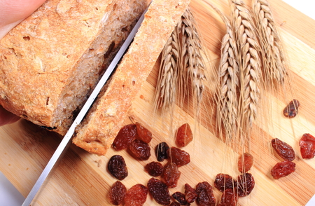 Slicing fresh baked wholemeal bread, ears of wheat and heap of raisins lying on cutting boardの写真素材