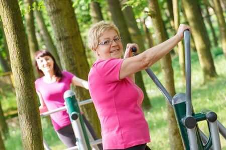 Elderly senior and young woman in sports wear exercising upper body on outdoor gym in sunny park, trainer machine, healthy, sporty lifestyles and slimming conceptの写真素材