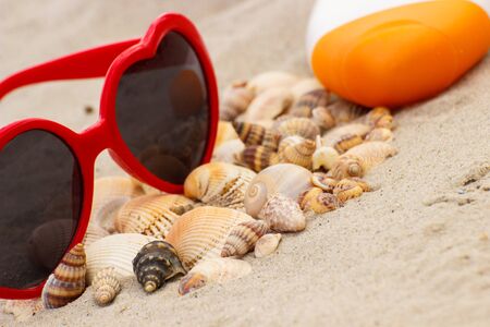 Heap of shells, red sunglasses in shape of heart and sun lotion on sand at the beach, seasonal concept, protection from sunの写真素材