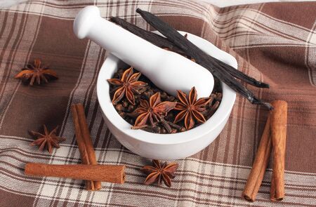 Fragrant cloves and anise in white glass mortar and spices on tablecloth on rustic wooden boardの写真素材