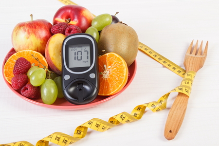 Glucose meter with result of sugar level, fresh fruits on plate and tape measure, concept of diabetes, slimming and healthy nutritionの写真素材