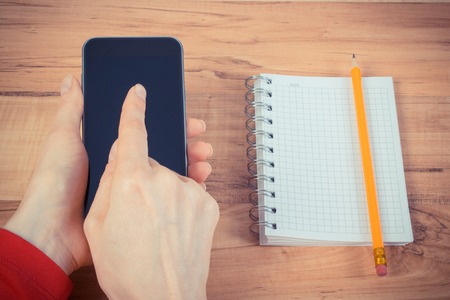 Vintage photo, Hand of woman touching blank screen of smart phone, notepad for writing notes in background, using mobile telephoneの写真素材