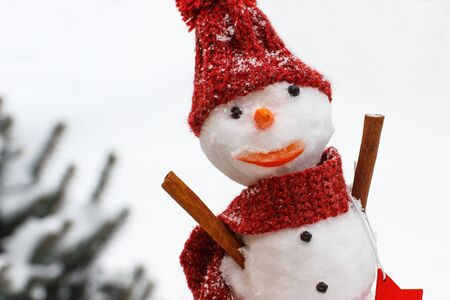 Funny snowman with woolen scarf and cap on background of coniferous tree covered snow, concept of winter seasonの写真素材