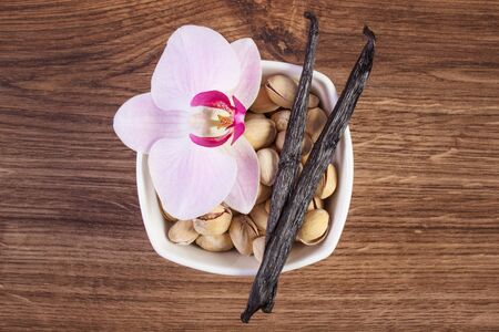 Pistachio nuts in white bowl, colorful blooming orchid flower and fresh fragrant vanilla sticks pods on wooden background, cosmetics ingredientsの写真素材