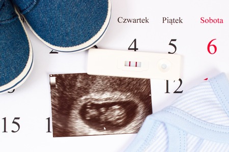 Pregnancy test with positive result, ultrasound scan of baby and clothing for newborn on calendar with polish inscription, concept of extending family and expecting for babyの写真素材