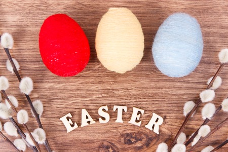 Inscription Easter, twigs of willow and colorful eggs wrapped woolen string on rustic board, festive...