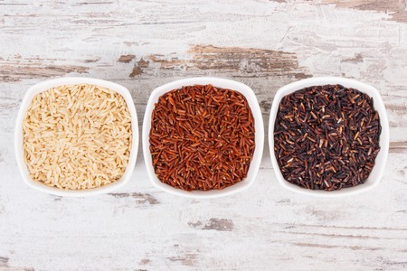 Black, red and brown rice in white glass bowls on rustic board, concept of healthy nutritionの写真素材