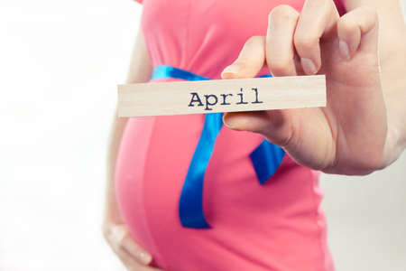 Woman in pregnant wearing pink dress with blue ribbon and showing word april written on wooden cube. Expecting for newborn and expansion of familyの写真素材