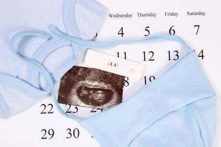Pregnancy test with positive result, ultrasound scan of baby and clothing for newborn on calendar, concept of extending family and expecting for babyの写真素材