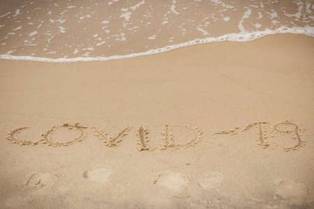 Inscription Covid-19 and incoming sea wave on sand at beach. Safe holidays during coronavirusの写真素材